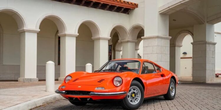 A classic Ferrari Dino is parked on a cobblestone driveway in front of a cream-colored building with arches and columns.