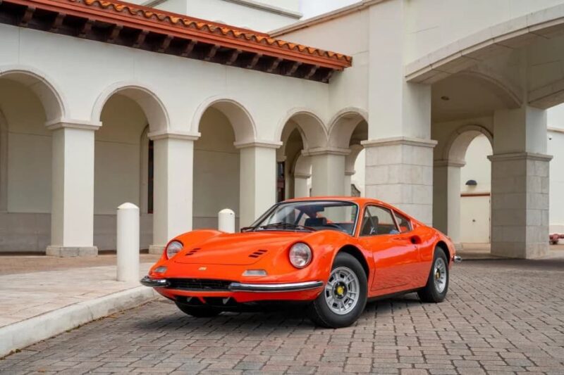 A classic Ferrari Dino is parked on a cobblestone driveway in front of a cream-colored building with arches and columns.