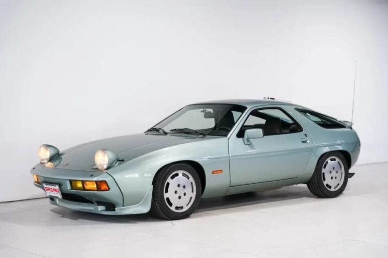A light green Porsche 928, an unsung classic from the Transaxle Porsche family, with pop-up headlights raised, is parked indoors against a plain white background.