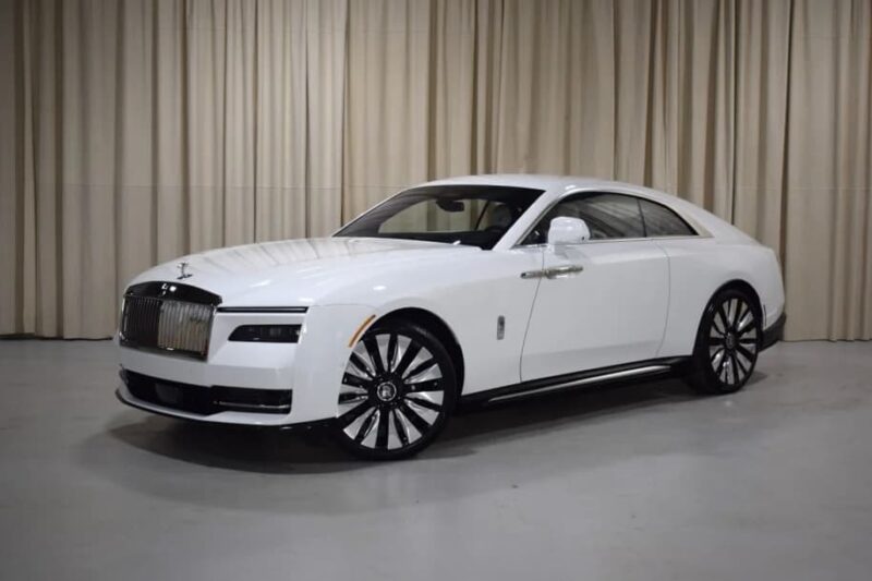 An ultra-luxe Rolls-Royce Spectre electric super coupe is parked indoors on a light-colored floor, framed by elegant beige curtains in the background.
