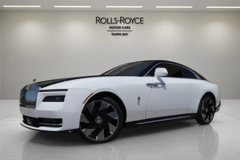 A white Rolls-Royce Spectre Electric Super Coupe is displayed in a showroom with "Rolls-Royce Motor Cars Tampa Bay" signage on the wall behind it.