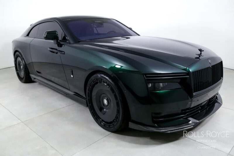 A dark green Rolls-Royce Spectre electric super coupe is parked in a white showroom, featuring tinted windows, black wheels, and a modern, angular design.
