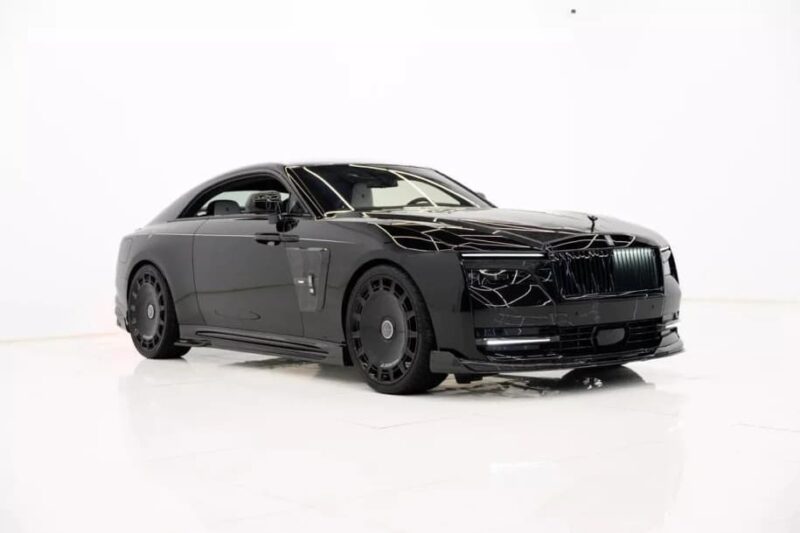 An ultra-luxe, black Rolls-Royce Spectre electric super coupe with tinted windows and custom rims is parked on a glossy white floor in a sophisticated studio setting.