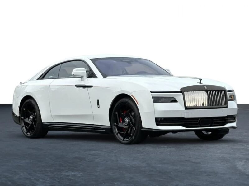 A white Rolls-Royce Spectre electric super coupe with black wheels and a prominent chrome grille, parked on a dark surface against a plain white background.