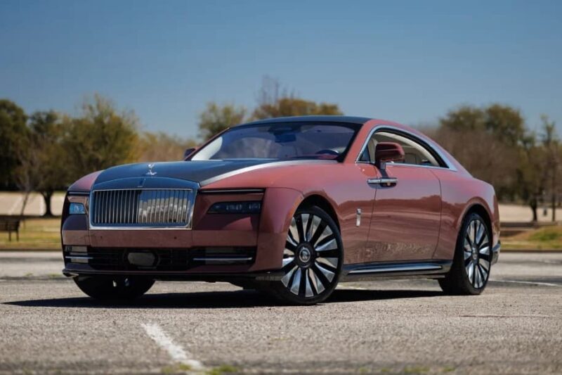 A maroon and silver Rolls-Royce Spectre electric super coupe is parked in an empty lot on a sunny day, with trees and a clear blue sky in the background.