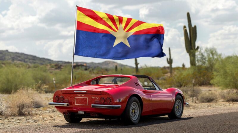 A sleek red sports car adorned with the Arizona state flag is parked on a desert road, cacti and mountains painting the backdrop. Perfect for any duPont REGISTRY feature, this beauty would steal the show at Arizona Car Week 2025.