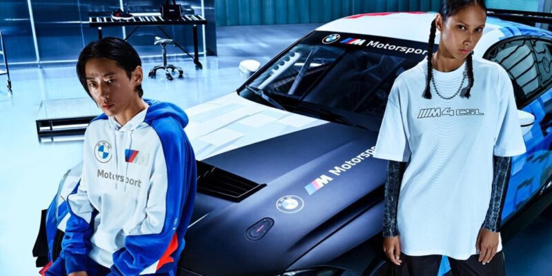 Two people in BMW Motorsport apparel stand next to a black BMW car adorned with racing decals, embodying a luxury lifestyle in a sleek, modern garage setting.