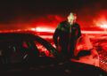 A person stands next to an open car door of a Porsche Taycan under red lighting. The scene is outdoors with snow on the ground, reminiscent of ice drifting, and blurred lights sparkle in the background.