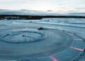 A Porsche Taycan gracefully glides on a snow-covered circular track with glowing red markers, mastering the art of ice drifting amidst a snowy landscape under a cloudy sky.