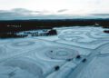 Aerial view of a snowy landscape featuring winding tracks and circular ice-drifting patterns, surrounded by trees and distant hills under a cloudy sky.