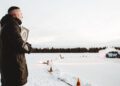 A person in a winter coat observes a Porsche Taycan effortlessly ice drifting on a snowy track, with trees in the background. Cones line the track, and smoke trails from the car's rear, echoing its recent Guinness World Record attempt.