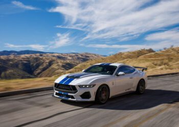 A white 2025 GT350 Shelby sports car with blue stripes roars along a mountain road under a clear, cloud-dotted sky. Its powerful 810HP engine takes on the curves effortlessly, with hills and valleys painting a stunning backdrop.