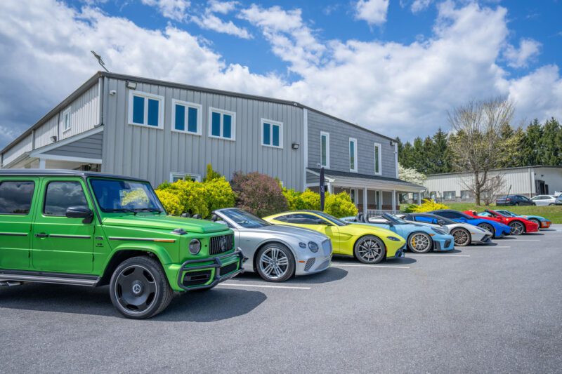 A row of luxury cars, courtesy of Magden Motors, including a green SUV and silver and yellow sports cars, parked in front of a modern building under a partly cloudy sky. Located in Jessup MD, this exclusive dealer showcases only the finest vehicles.