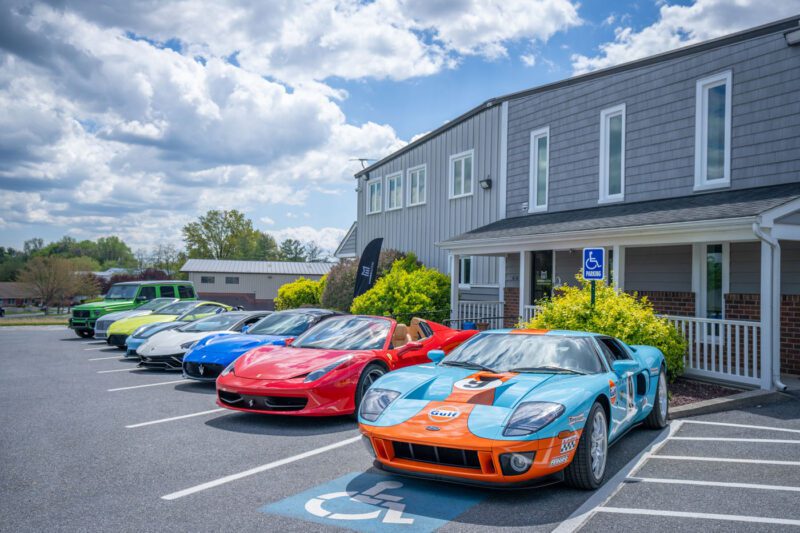 A row of six luxury sports cars is parked in front of a building, including a blue and orange Ford GT in a handicapped space. Underneath the partly cloudy sky, these vehicles shine with elegance thanks to Magden Motors, an exclusive dealer known for their premium selection near Jessup MD.