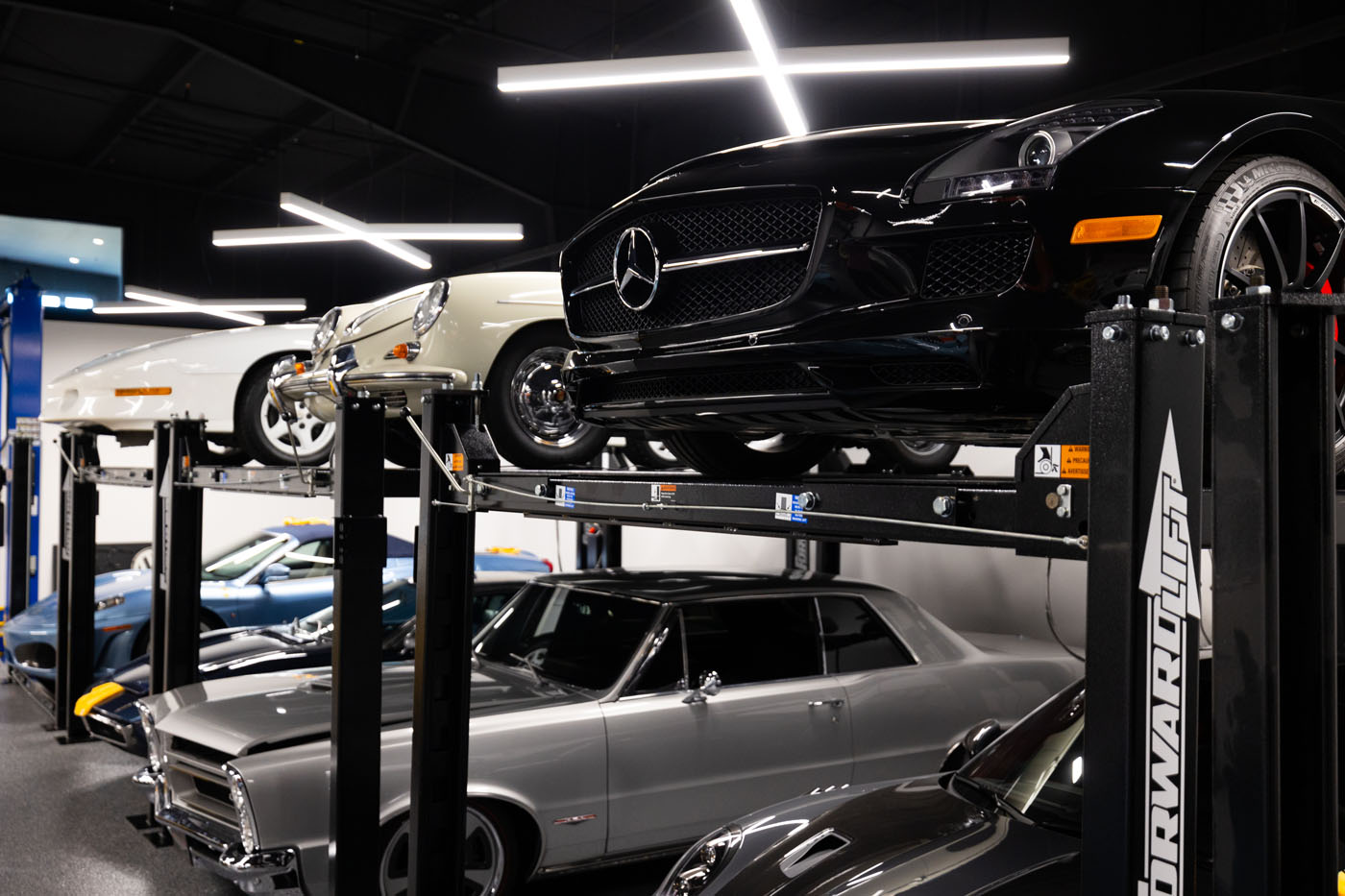 A row of vintage and modern cars is displayed on lifts in the Magden Motors showroom in Jessup, MD, illuminated by fluorescent lights.