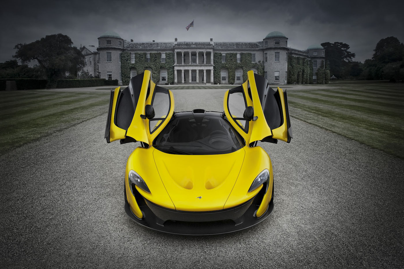 A McLaren P1 with its scissor doors open is parked on a gravel path in front of a large, historic mansion under a cloudy sky.