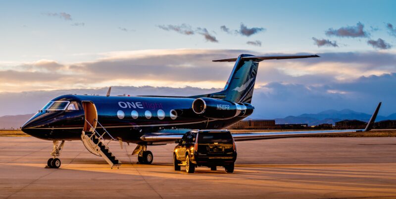 A private jet with "ONEflight" branding is parked on a runway adjacent to a black SUV, silhouetted against a sunset sky. This elegant scene captures the essence of luxury travel as seen in the 2025 duPont REGISTRY's Aviation Guide, highlighting opulence and seamless style.