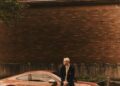 A man in a blazer and plaid pants leans against a Porsche, part of Poggy's exclusive capsule collection, parked on a street with a brick wall and lush greenery in the background.