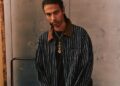 A person with braided hair wearing a striped jacket from the Poggy Capsule Collection stands indoors near stacked tires, evoking a sleek Porsche aesthetic.