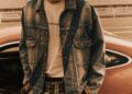 A person with blond hair stands beside a parked Porsche, wearing a denim jacket, white shirt, and plaid pants from the Poggy Capsule Collection.
