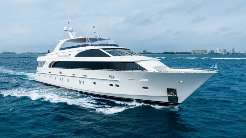 A large luxury yacht sails on open water under a cloudy sky, with a distant shoreline and buildings visible in the background, offering an exquisite maritime experience.