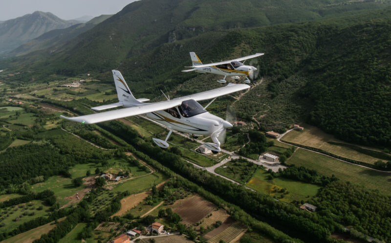 Two small white airplanes fly in formation over a lush, green landscape with fields, trees, and winding roads, set against distant hills—an aerial spectacle worthy of the 2025 duPont REGISTRY Aviation Guide.