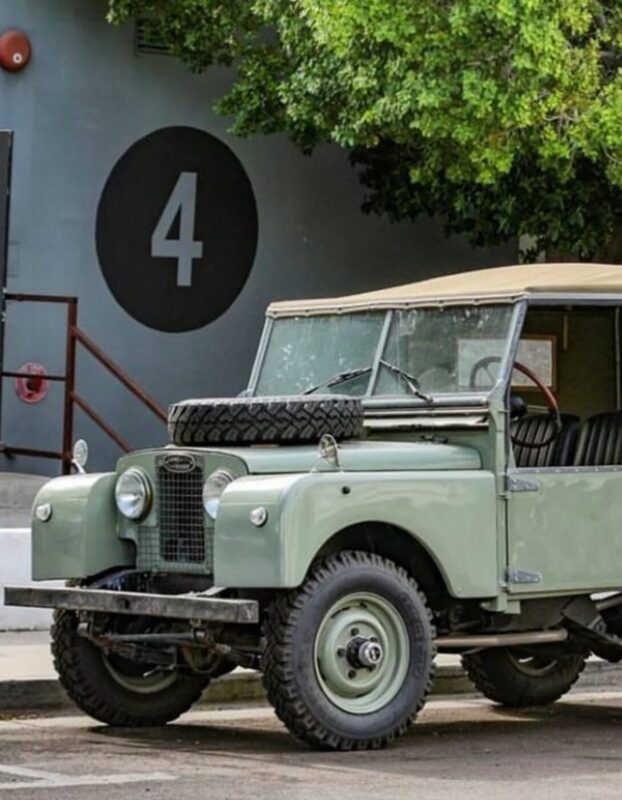 A vintage light green off-road vehicle with a spare tire on the hood is parked near a building adorned with a large number 4, reminiscent of the adventures highlighted in the duPont REGISTRY car guide.