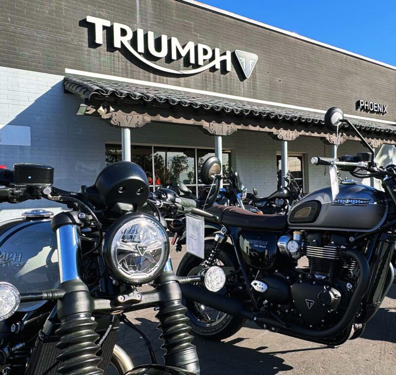 Two Triumph motorcycles parked in front of a Triumph dealership, capturing the essence of Arizona Car Week 2025. This moment, worthy of the duPont REGISTRY spotlight, showcases the allure and craftsmanship synonymous with the brand amidst an event celebrating automotive excellence.