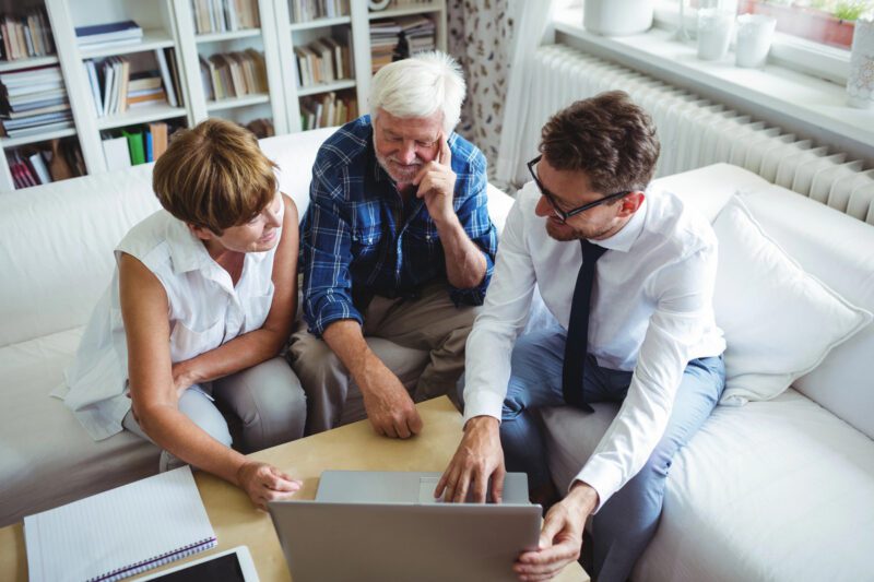 Three people sitting on a couch intently examine a laptop screen, perhaps discussing an estate settlement. A notebook and pen lie on the table, ready for personal process notes. Shelves filled with books provide a thoughtful backdrop to their focused conversation.