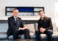 Two men sitting on a gray sofa in a modern room; one wears a suit and tie, the other, possibly Bruno Spengler, sports a black jacket with the subtle flair of a Pilote Officiel.