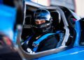 Race car driver and Pilote Officiel, Bruno Spengler, wearing a black helmet and safety harness, sits inside the blue cockpit of his Bugatti vehicle, looking confidently towards the camera.