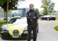 A man in a black jacket stands confidently, arms crossed, as Bruno Spengler—a Pilote Officiel—might, in front of a yellow and black Bugatti on a gravel driveway. Another Bugatti gleams in the background near a sleek modern building.