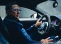 A man wearing glasses and a padded vest is sitting in the driver's seat of a Bugatti, holding the steering wheel with the poise of a Pilote Officiel.