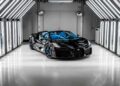 The sleek Bugatti sports car, with its black finish and striking blue accents, basks under geometric light panels in a well-lit showroom. The unveiling of the W16 Mistral adds an air of excitement to the scene.