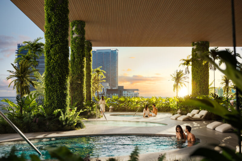 The rooftop pool area at Tower 2 boasts green walls, lounge chairs, and a serene ambiance as several people enjoy the pools at sunset. Tall buildings and palm trees accentuate the skyline, highlighting Miami Real Estate's vibrant allure.