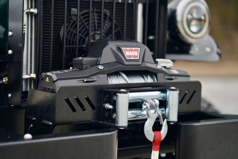 A close-up of a front-mounted electric winch on a Timberline Green Power Wagon. The winch, exuding modern luxury, features a cable and hook with a visible red warning label.