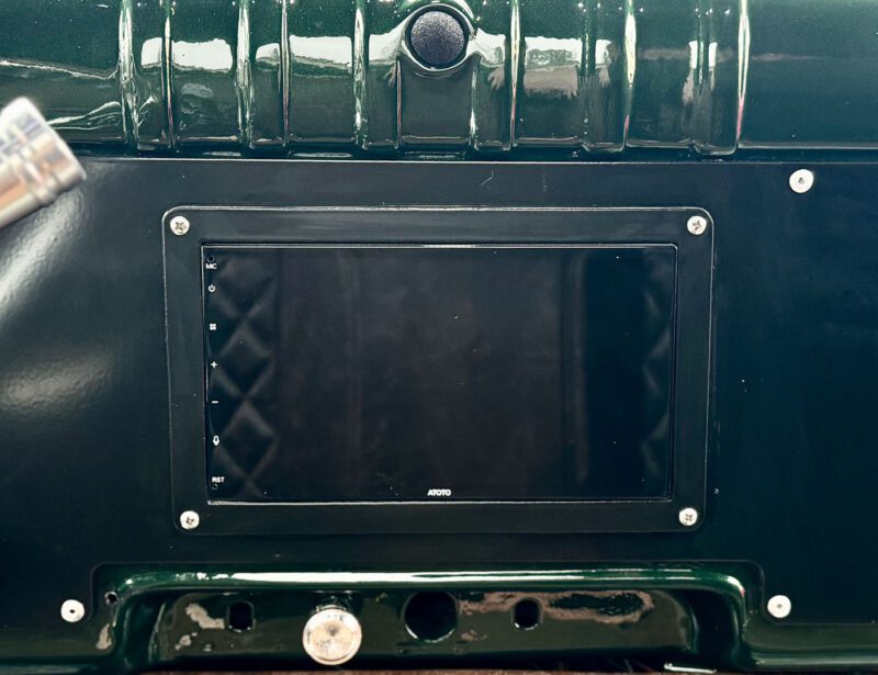 Close-up of a car dashboard featuring a rectangular screen surrounded by buttons and screws, all showcasing the sleek modern luxury of a dark 1946 Timberline Green finish.