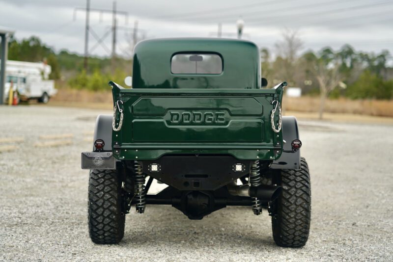 Rear view of a dark green vintage Dodge Power Wagon, exuding timeless power on gravel. Its raised suspension and large off-road tires hint at its resilience, embodying the essence of a true desert power wagon ready for any adventure.