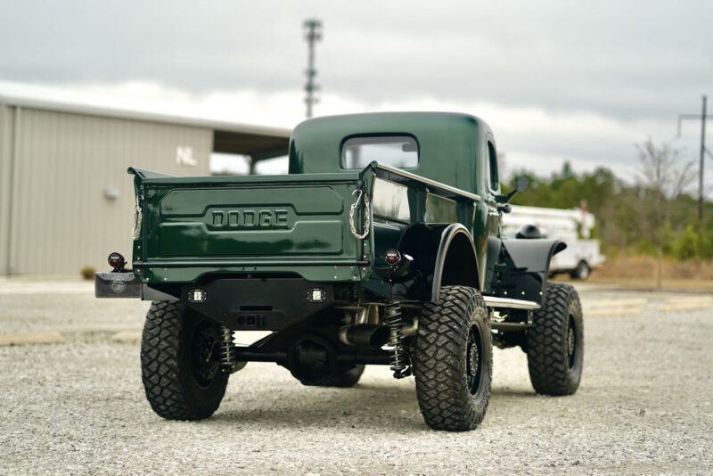 A Timberline Green vintage Dodge Power Wagon with large off-road tires rests on gravel near a building, combining classic charm with a hint of modern luxury.