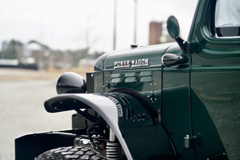 Close-up of the front part of a dark green vintage Desert Power Wagon, showcasing its headlight, fender, and the iconic "Power Wagon" emblem on the side. The blurred outdoor background accentuates its timeless power.