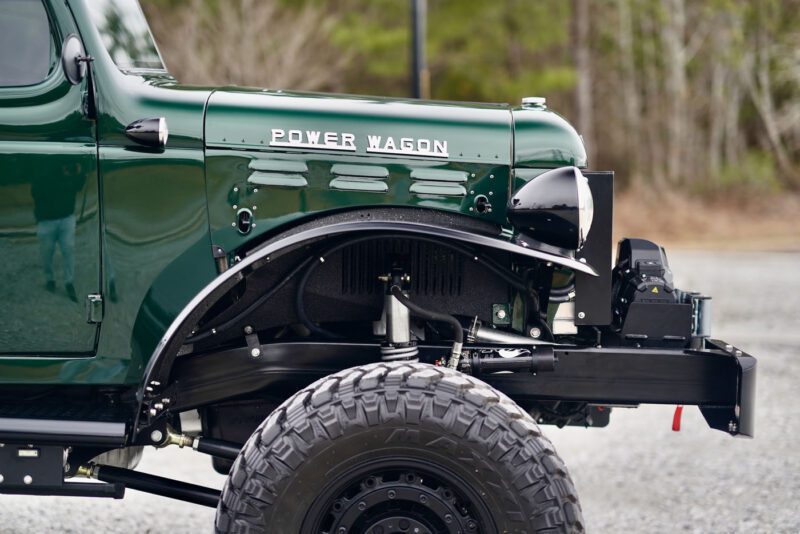 Close-up of a vintage Timberline Green Desert Power Wagon truck, front fender removed to reveal the intricate suspension and engine components, set against a beautifully blurred outdoor backdrop.