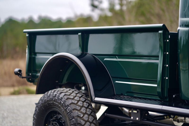 Close-up of a green truck bed with black fender, parked on a gravel surface. The rugged design evokes the spirit of a Desert Power Wagon. Trees are blurred in the background, adding a touch of timeless power to this scene.