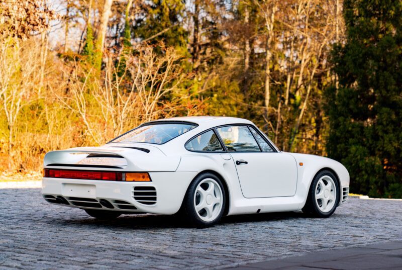 A white Porsche 959, renowned and rare as featured in duPont REGISTRY, stands elegantly parked on a cobblestone surface, its sleek lines accentuated by the surrounding trees.