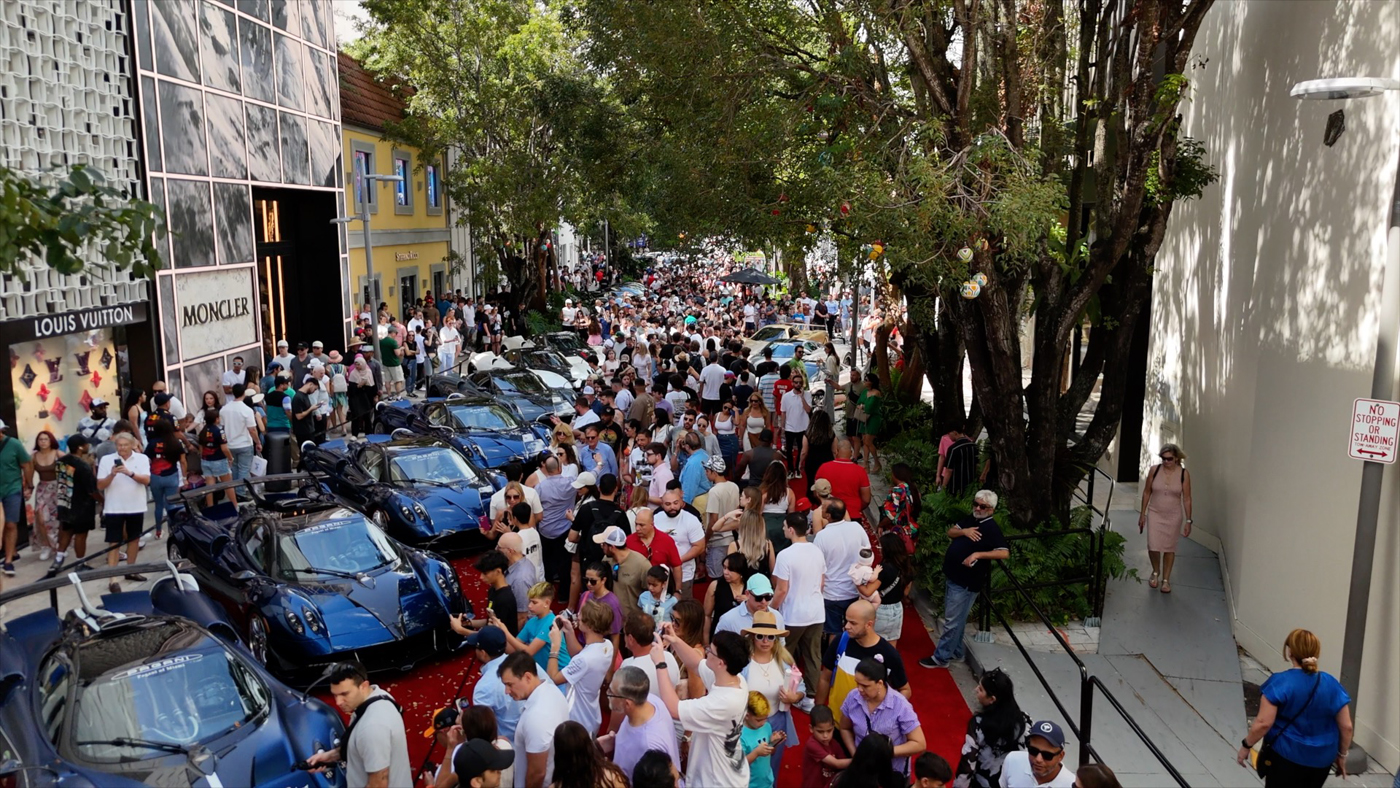 A large crowd gathers at the 2025 Miami Concours, an exquisite Supercar Art Event. Numerous people admire the lineup of parked sports cars along the tree-lined street, where innovation meets elegance under the sun.