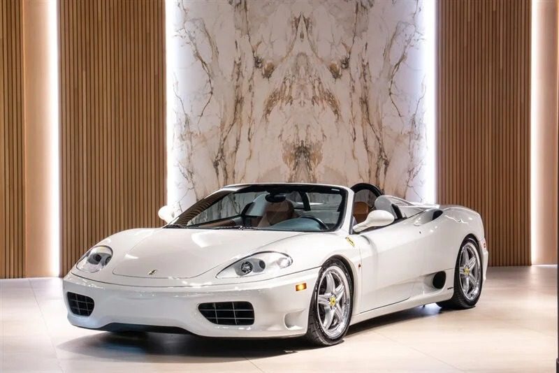A white convertible sports car from the 2000s is parked indoors on a light-colored floor, exuding nostalgia against a marble-patterned wall and vertical wooden panels in the background.