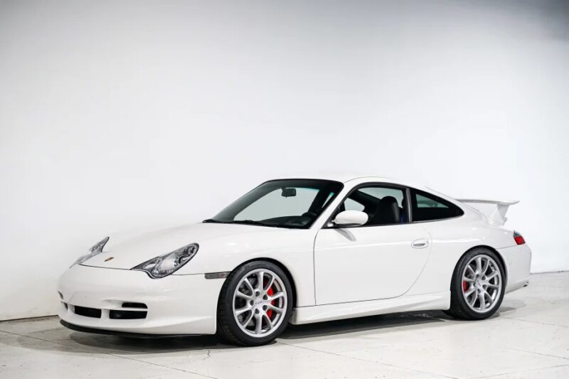 A white sports car with a sleek design and rear spoiler, reminiscent of the best cars from the 2000s, is parked indoors against a plain white wall.