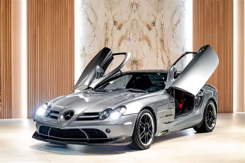 A sleek silver sports car with gullwing doors open, reminiscent of iconic 2000s cars, is parked indoors on a polished white floor. The background features vertical wooden panels and a marble accent wall. This stunning vehicle is now available for sale, inviting both nostalgia and admiration.