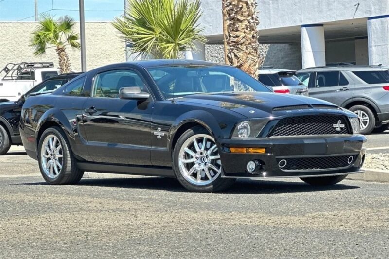 A nostalgic black sports car from the 2000s sits parked outdoors, showcasing alloy wheels and a distinctive front grille. Other vehicles and palm trees frame this timeless beauty—perhaps it's even for sale?.