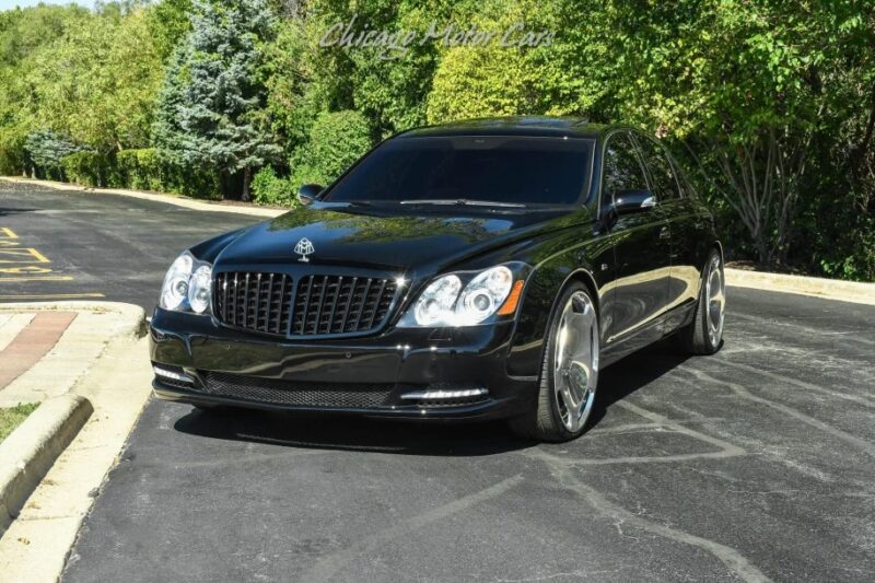 A black luxury sedan, reminiscent of the best cars from the 2000s, is parked on a sunlit road surrounded by lush greenery.