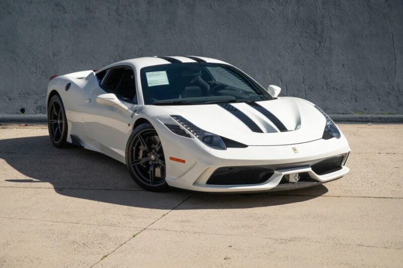 A 458 Speciale, one of the best modern Ferraris, sits gracefully with its black racing stripes cutting across its white form, parked on a concrete surface against a plain gray wall.